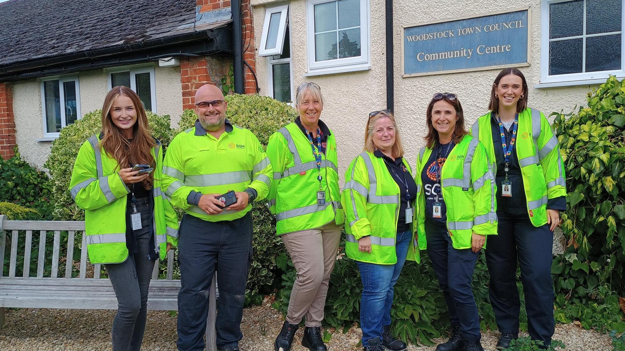 Four women and a man in SGN branded hi viz yellow jackets are smiling for the camera in front of Woodstock Town Council Community Centre. A bench and some greenery could be seen behind them.