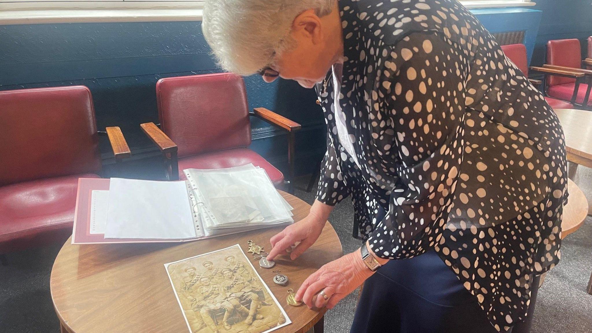 A woman with grey hair bends to look at a photo and some medals set out on a round wooden table.