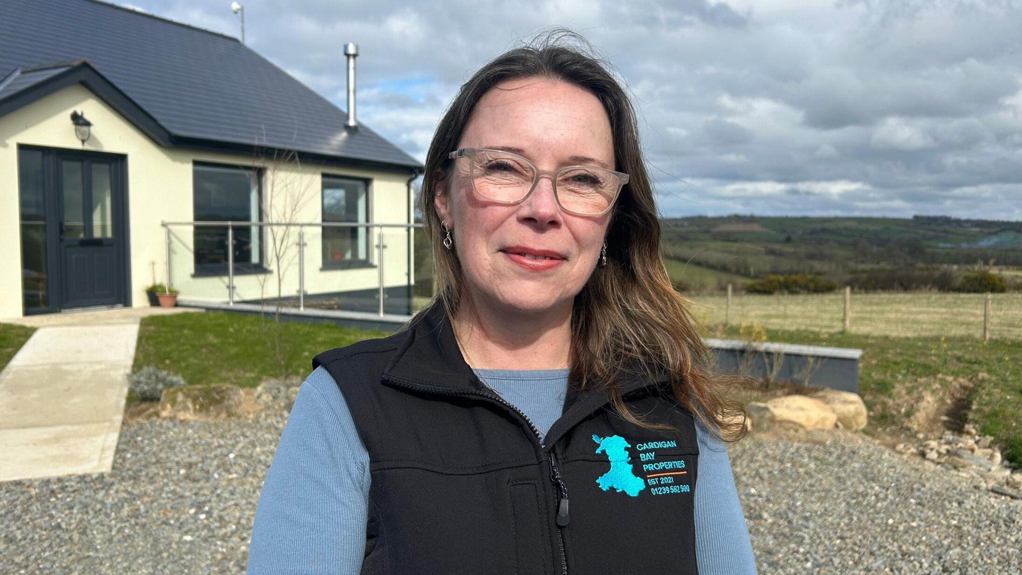 Tania Dutnell, estate agent. Tania has long brown hair reaching to her shoulders and has glasses and silver dangling earrings. She is wearing a light blue long sleeved top with a black bodywarmer on top. The bodywarmer has a logo with a light blue map of Wales and the words 'Cardigan Bay properties'. Behind her is a cream house with black doors and fields behind. 