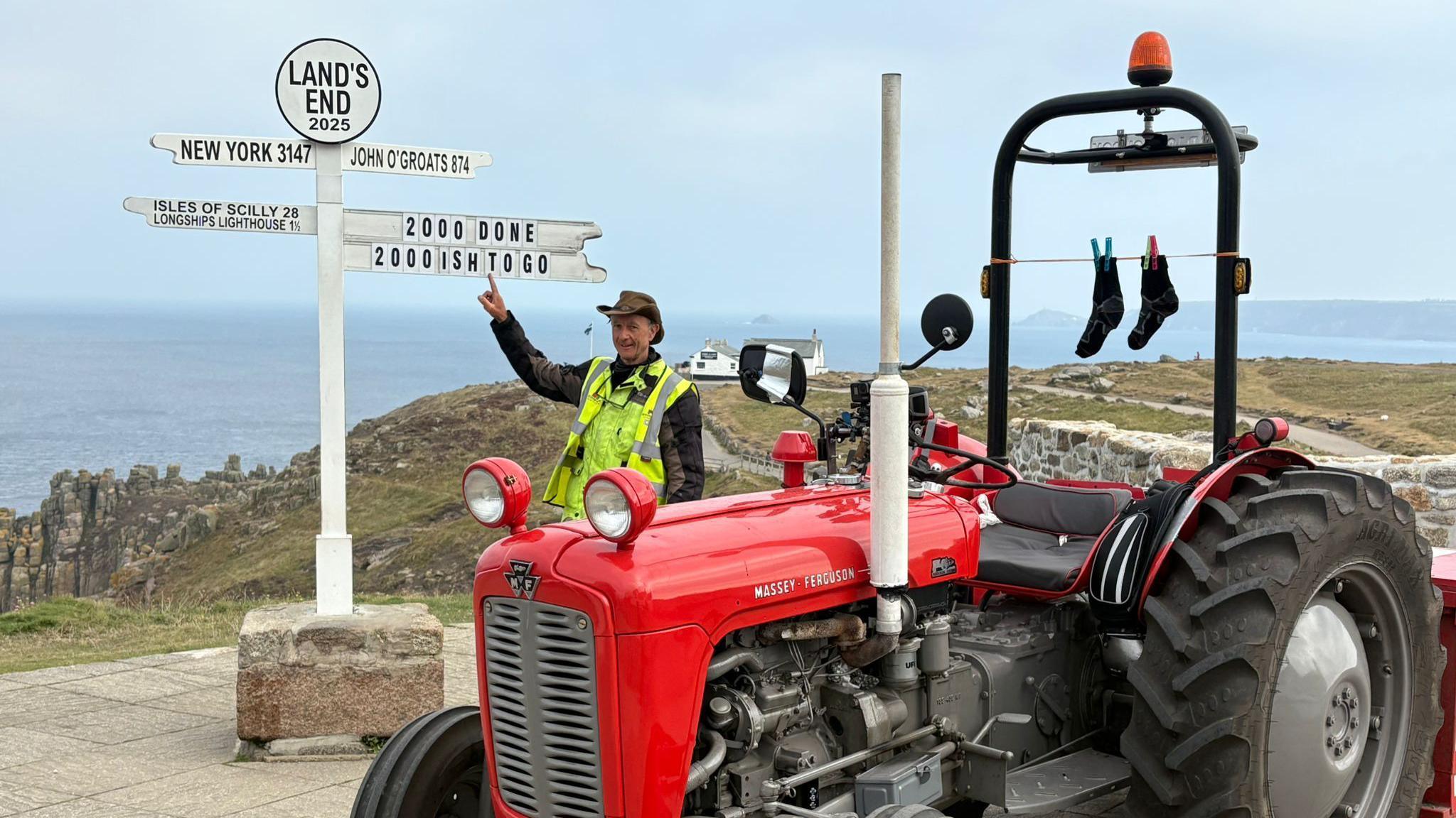 Marius Anderson stood being a red Massey Ferguson 35x tractor. He is pointing to the Lands End Sign. The Sign has been modified to say "2000 done 2000 ish to go." The sky is grey. The sea is a light blue.
