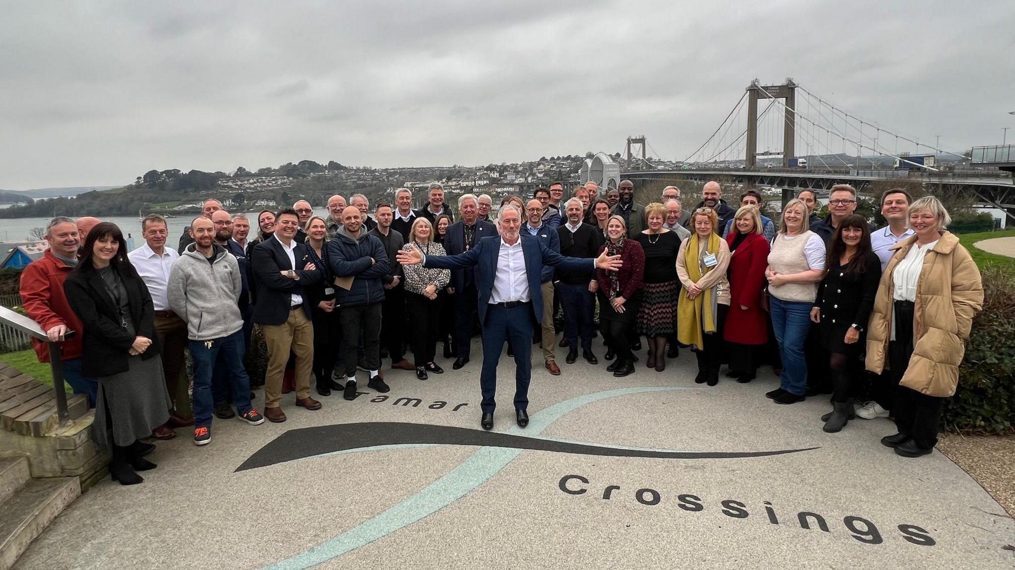 David List stands in front of a group of colleagues during a party outside near the Tamar Bridge on the Plymouth side of the river to mark his retirement. It is a cloudy day and the Tamar Bridge can be seen in the background.