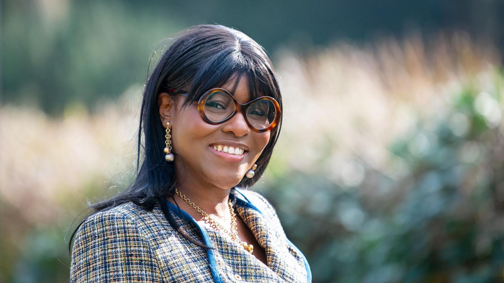 Onike Gollo wearing round glasses and a blazer while smiling at the camera. She has a full fringe and is wearing earrings