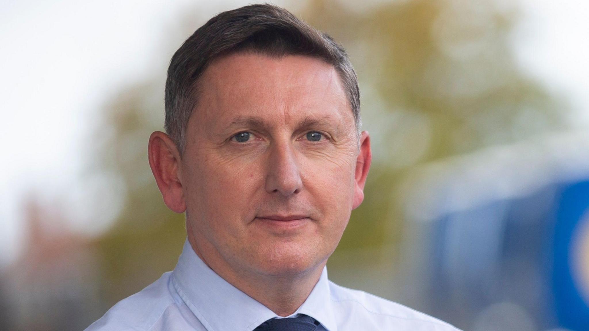 Steve Sheridan wearing a white shirt and dark tie. He has short brown hair. 