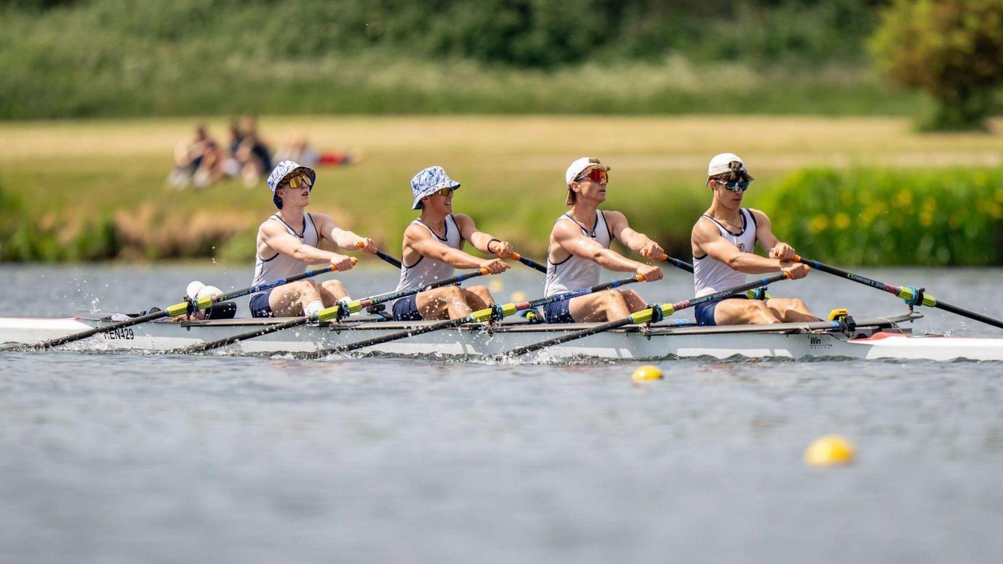 Four young men row a boat mid-competition on a river. They wear white vests, blue shorts, sunglasses and sunhats.
