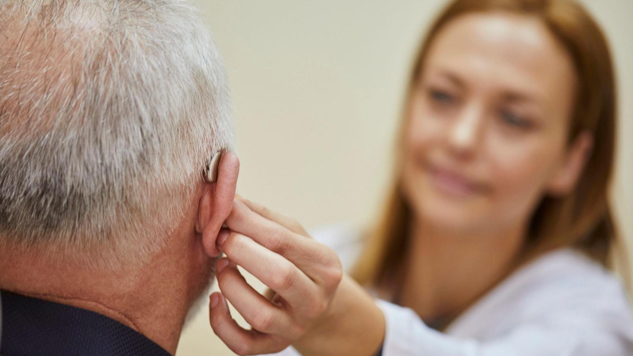 A close-up view of an older individual with short, grey hair, seen from behind. The person is wearing a hearing aid in their right ear. A female health professional, whose face is out of focus in the background, is gently adjusting or fitting the hearing aid, with her hand placed on the older person's ear. The background is softly blurred.
