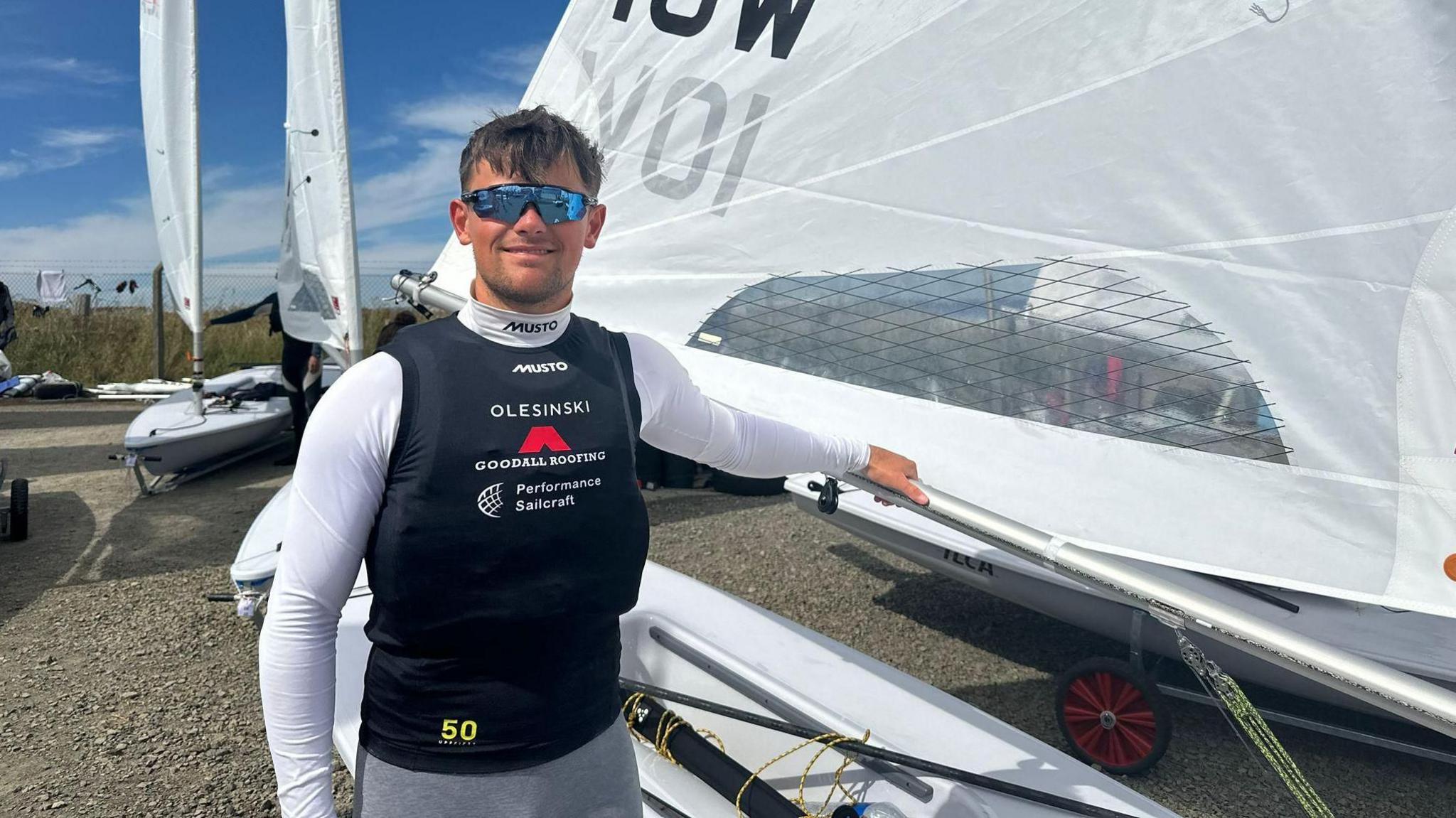 A smiling Arthur wearing light blue reflective sunglasses, a black waterproof vest with a white long sleeved wetsuit. He has placed his left arm on a boat.