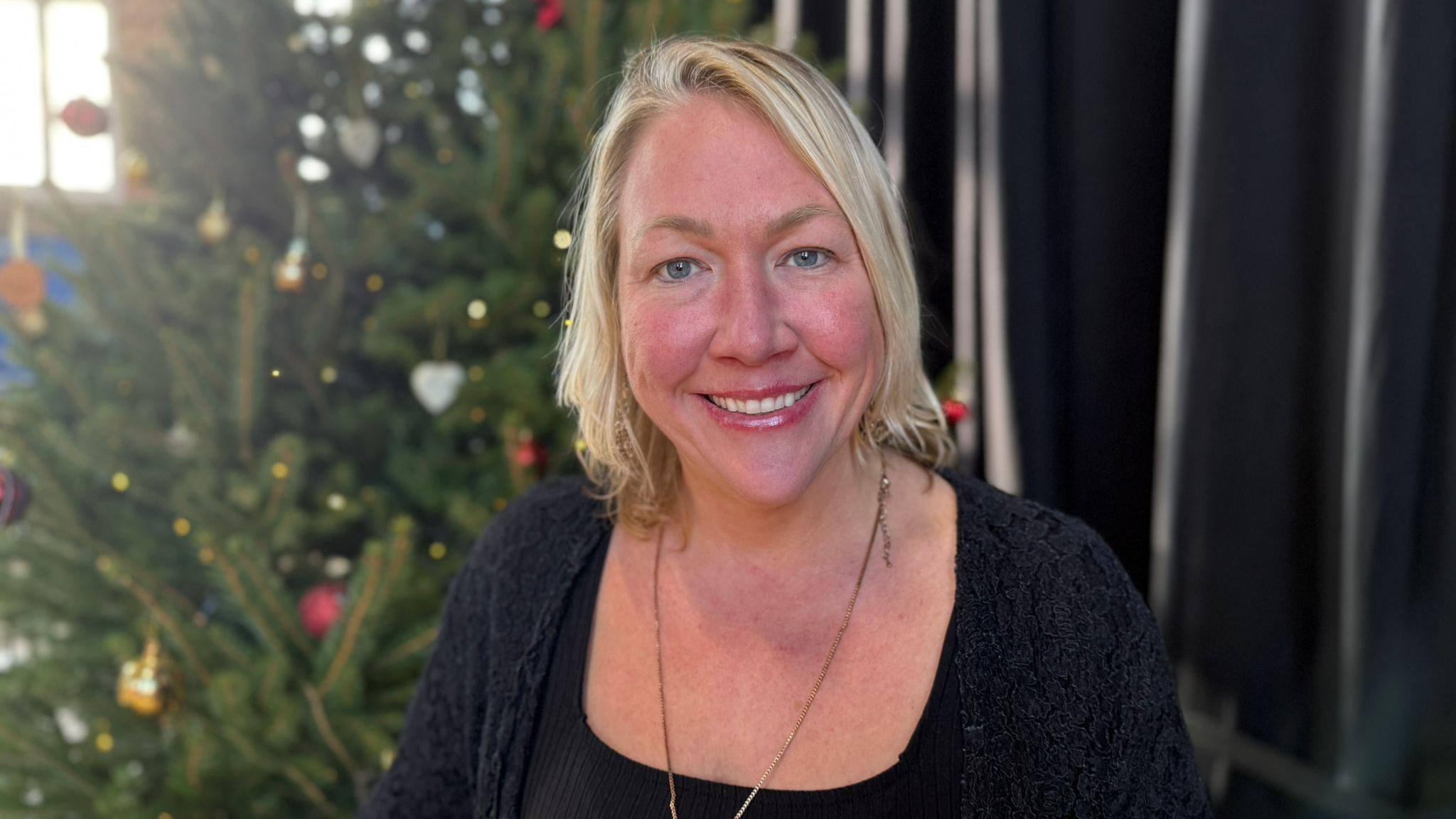 A woman with blonde hair smiles at the camera with a Christmas tree behind