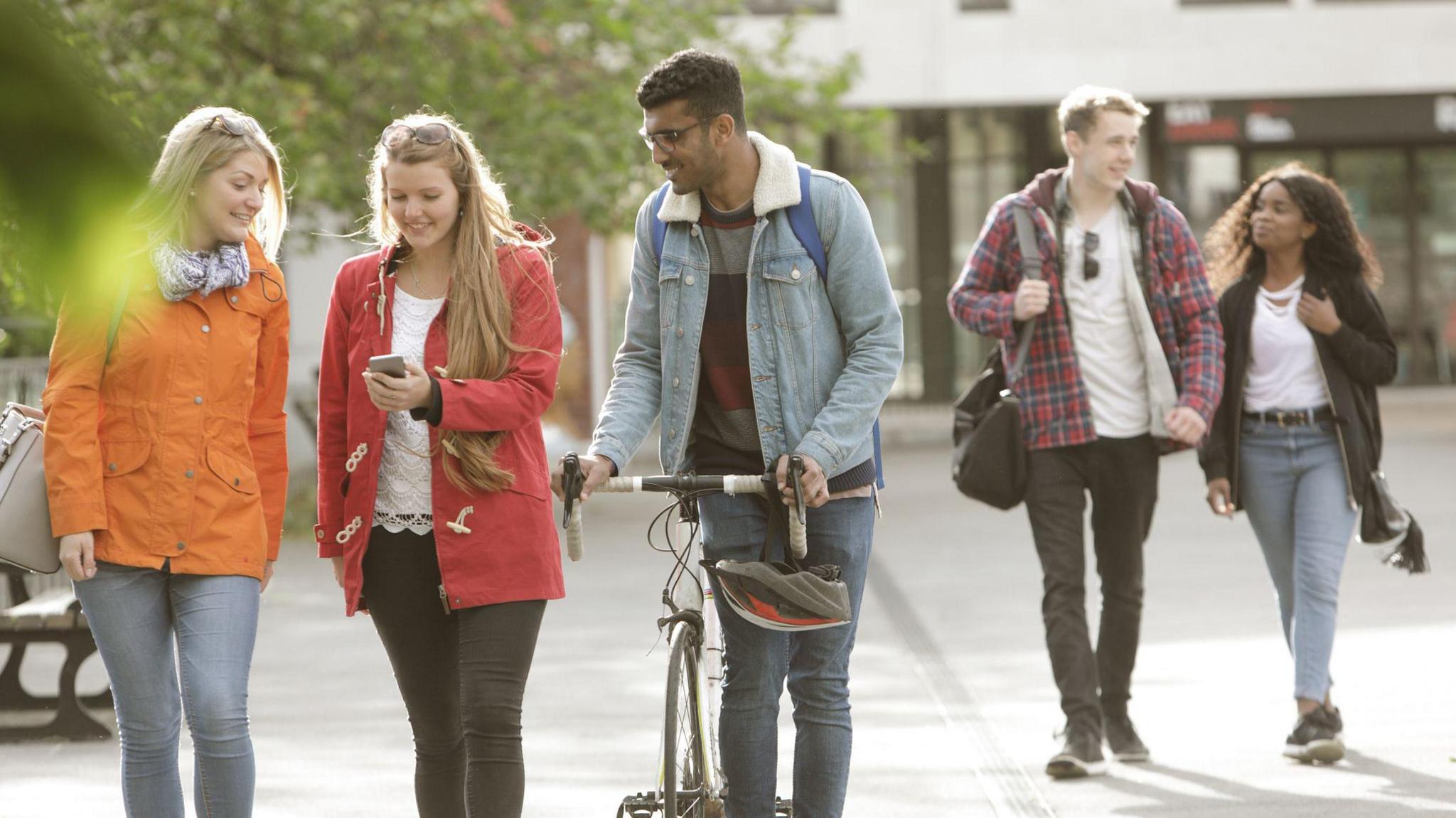 A group of students walk outside a campus building. In the foreground, one female student is showing her phone to another female student and a male student pushing a bicycle. In the background, a male and female student walk together separate from the other group.