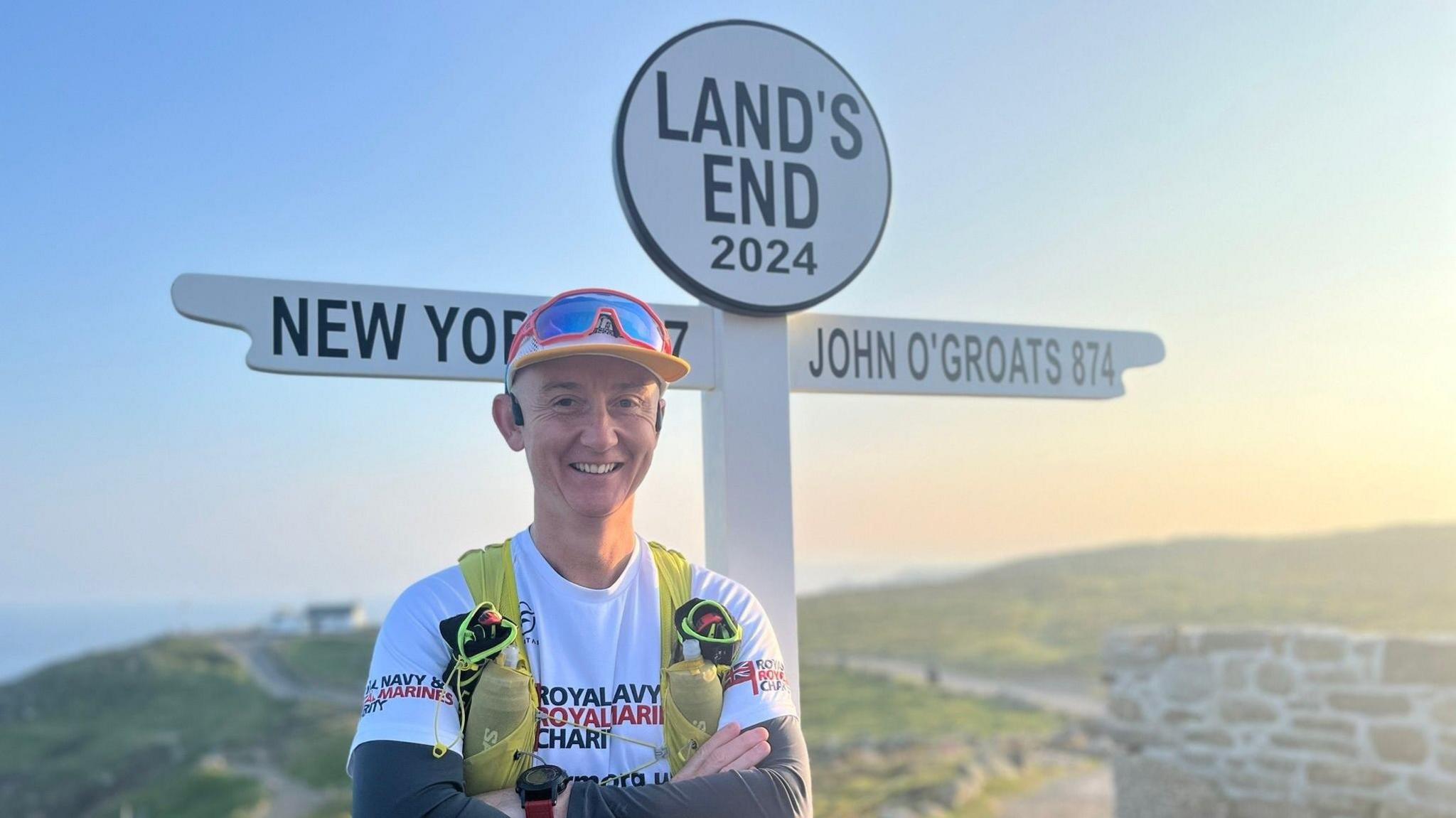 Alex Mills, a man smiling and wearing a cap with goggles, stands next to a sign which reads New York, Land's End and John O'Groats