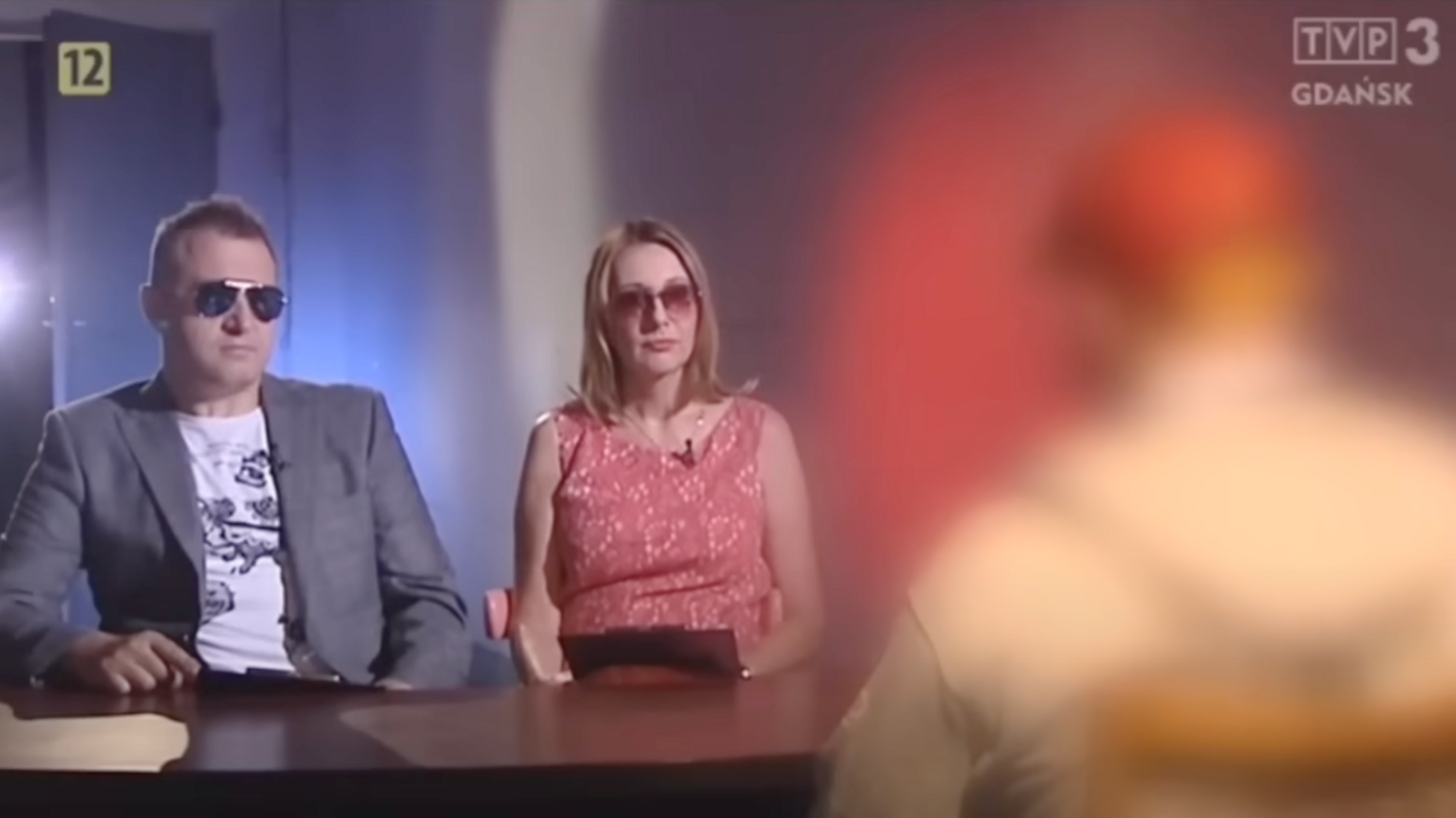 Two Polish interviewers wear sunglasses while sitting across a table from a blurred-out man.