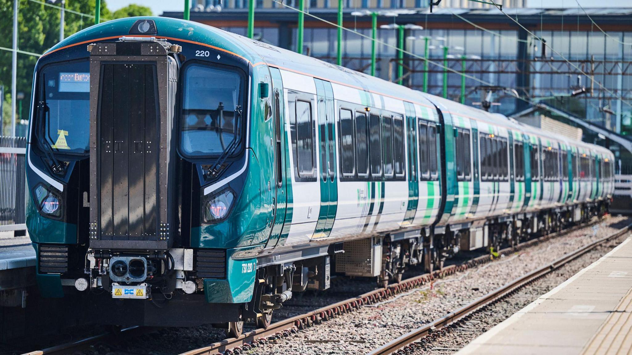 A new Class 730/2 training, pulling out of a railway station, with a building behind. The train has a large number of carriages and is green and white in colour.