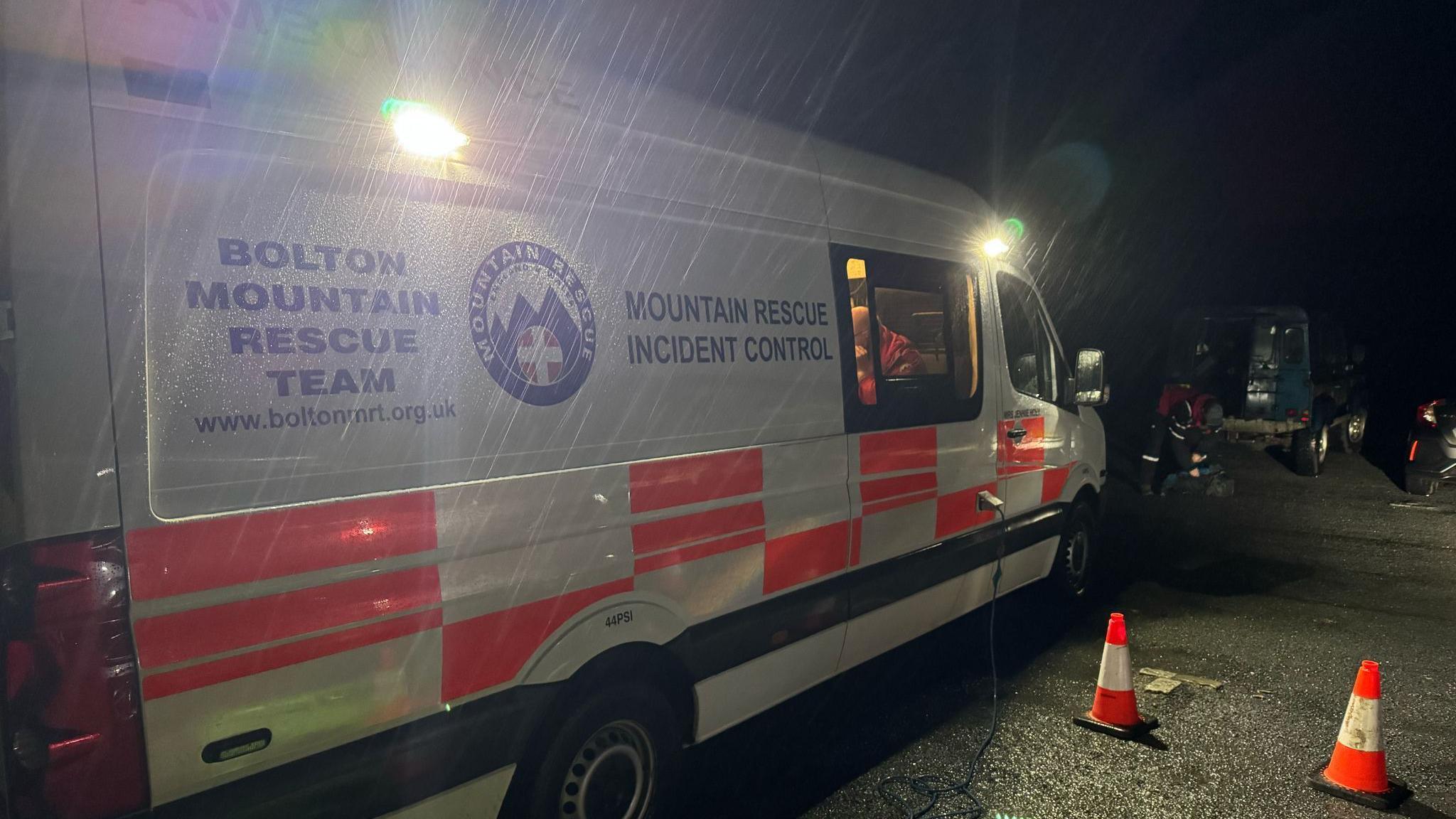 A mountain rescue vehicle parked by some traffic cones in the dark at a car park. Rain can be seen lashing against the side of the vehicle. 