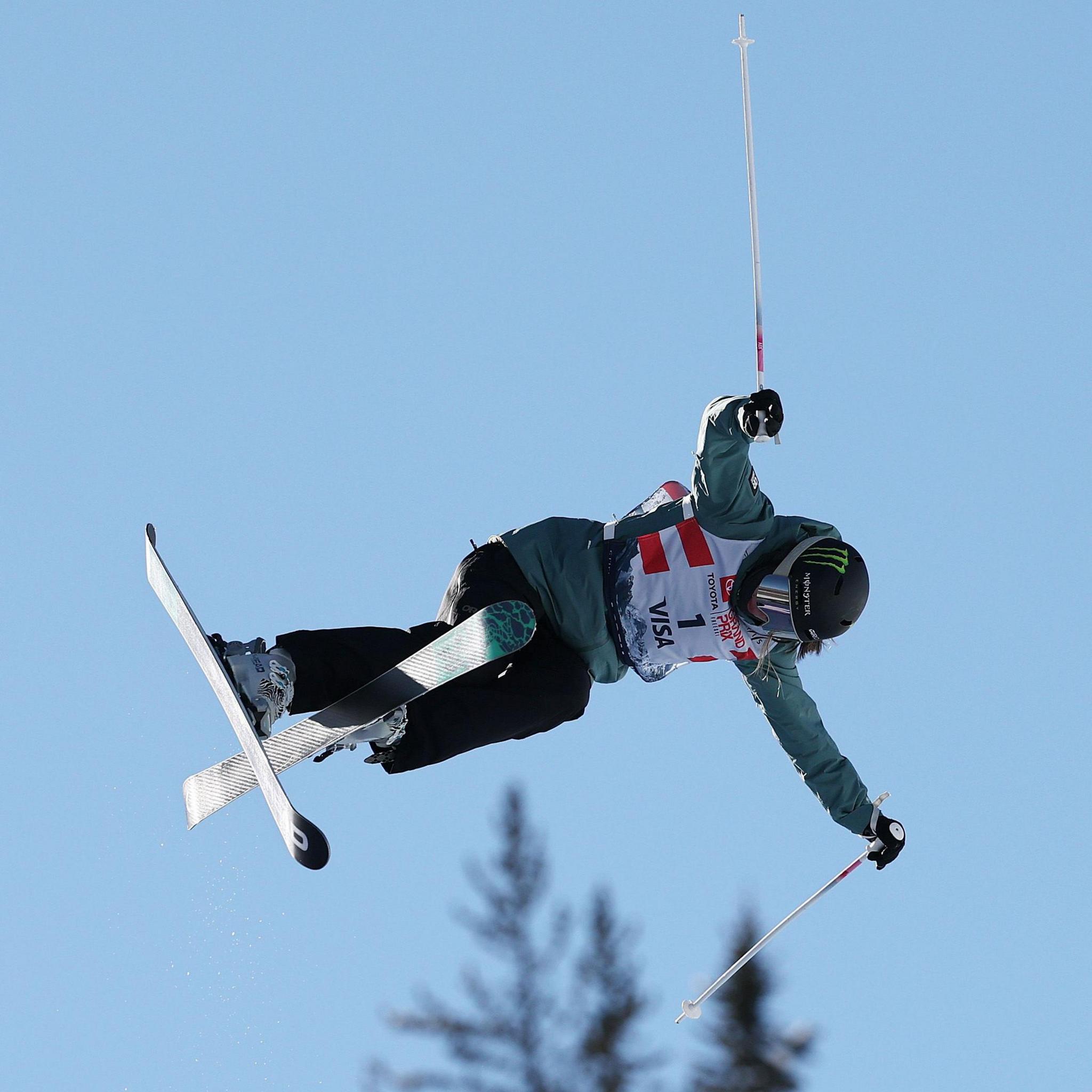 Freestyle skier Zoe Atkin is airborne, with only a blue sky and the tops of trees visible behind her
