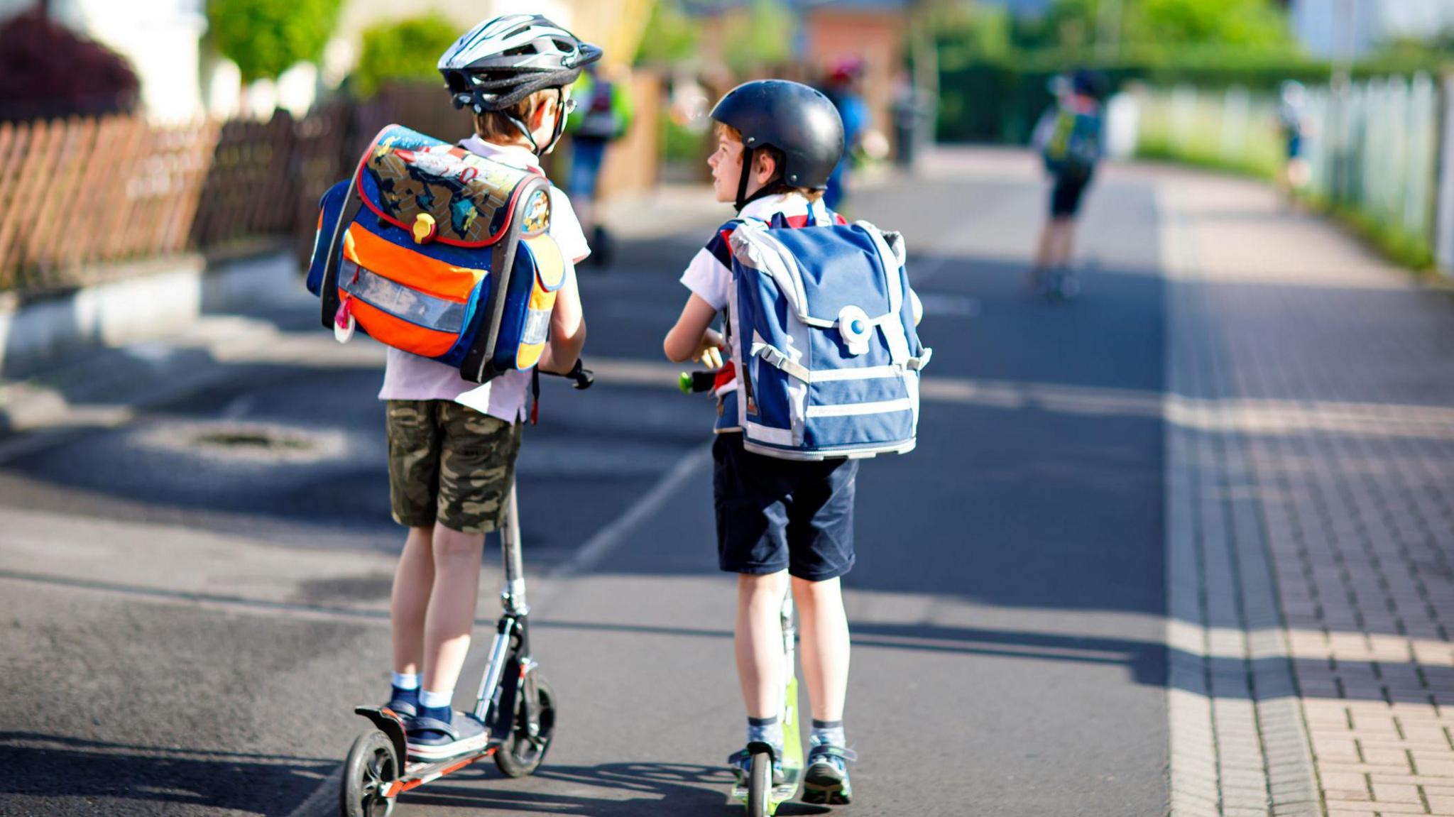 Two young boys on scooters wearing backpacks leaving school in hot sunny weather.