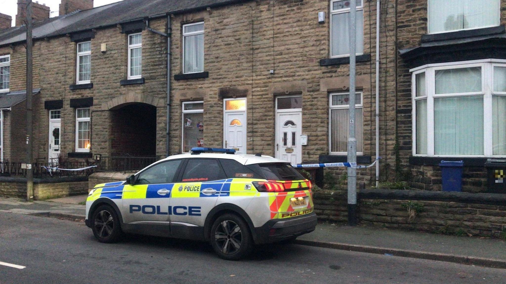 A police car is parked outside a row of terraced houses. One of the houses has blue and white police tape strung outside.
