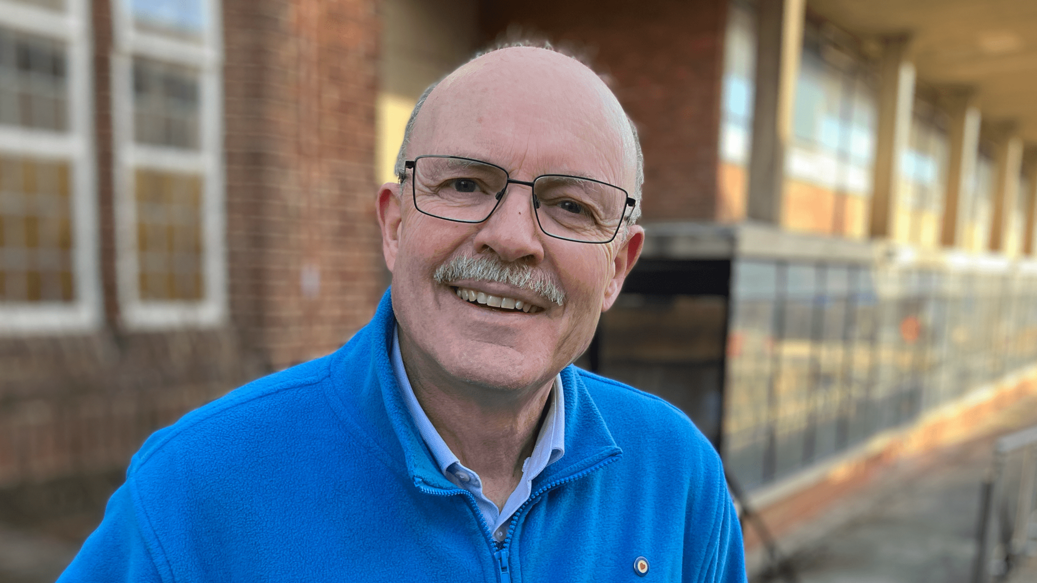 Alan Matlock has short grey hair, a grey moustache and thin black glasses. He is wearing a blue fleece, and an unidentifiable building is out of focus behind him.