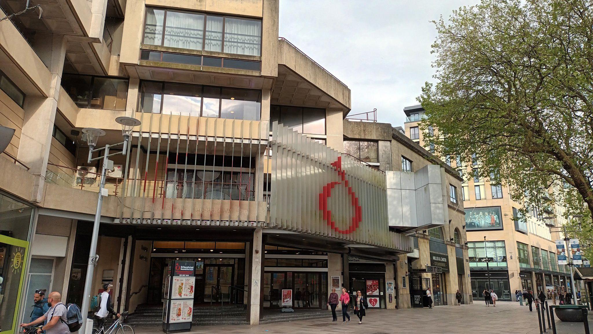 St David's Hall in Cardiff - a large brown building with a red letter D on top and windows, and people walk past the doors 