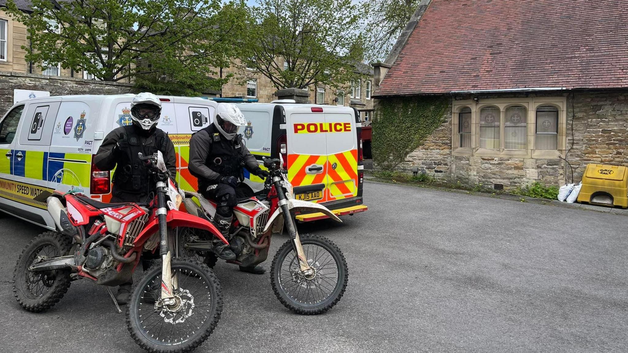 Two people dressed in black with helmets standing behind motorcycles. They're in front of two police vans.