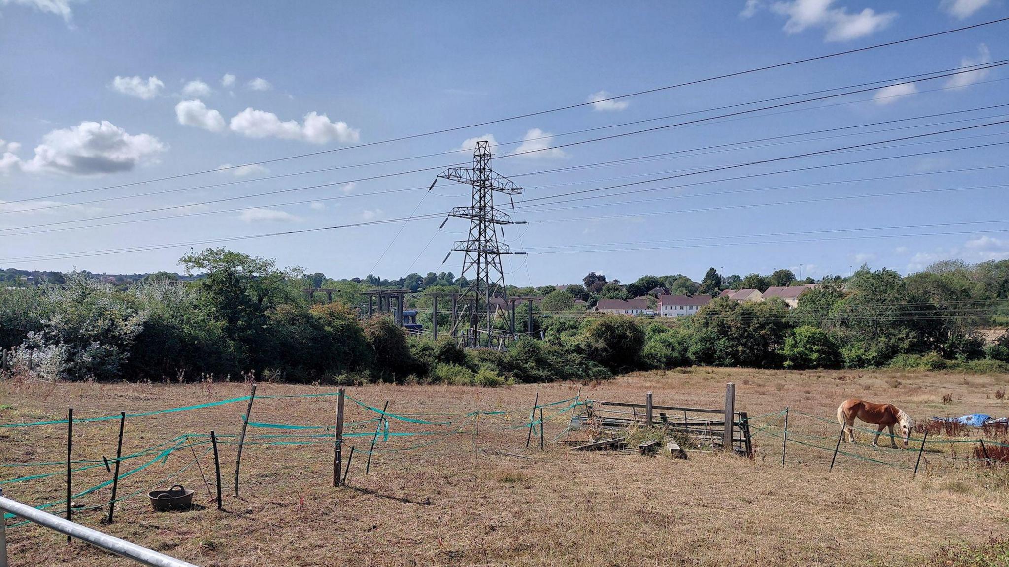 A brown field, with some green ribboned fencing inside. A horse is wondering the paddock on the right hand side of the frame. In the distance are green trees, houses and an electricity pylon.