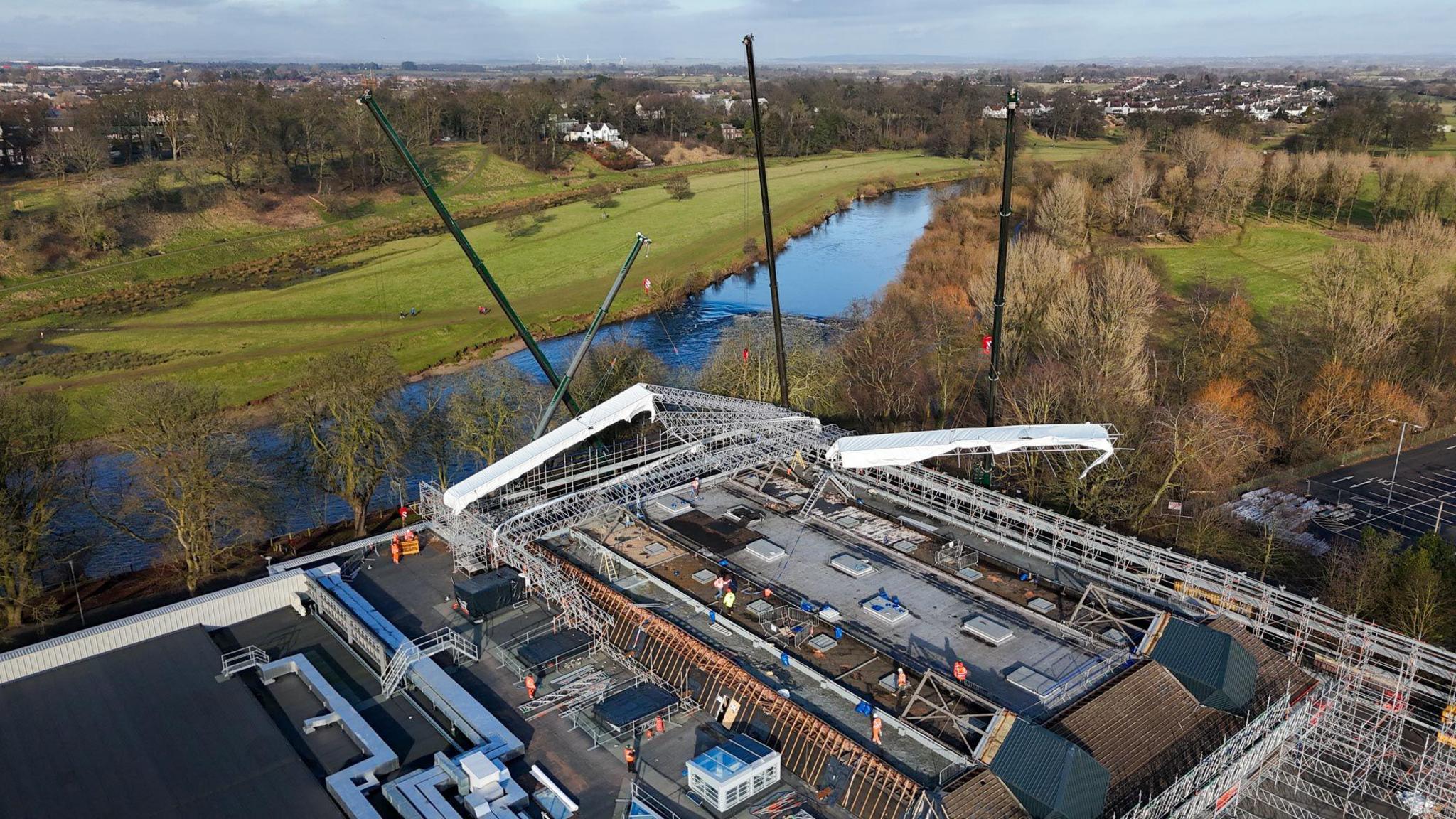 An aerial photo of the Sands Centre roof which is partially covered with silver scaffolding. A river and fields can be seen beyond the building.