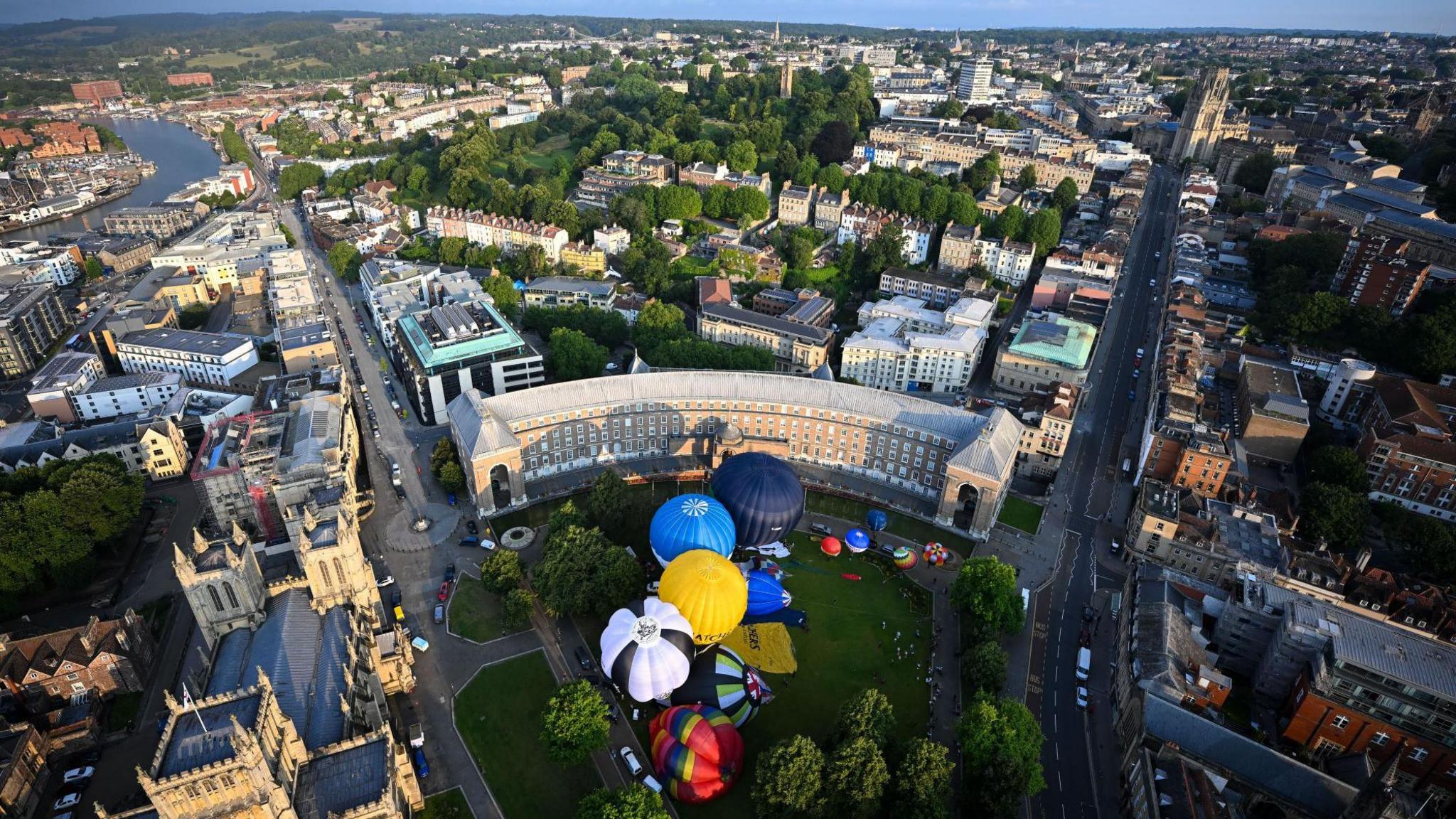 A cluster of colourful hot air balloons are seen from the air on College Green in the centre of Bristol. The image also shows a large part of the city in the background including Park Street and the harbourside.