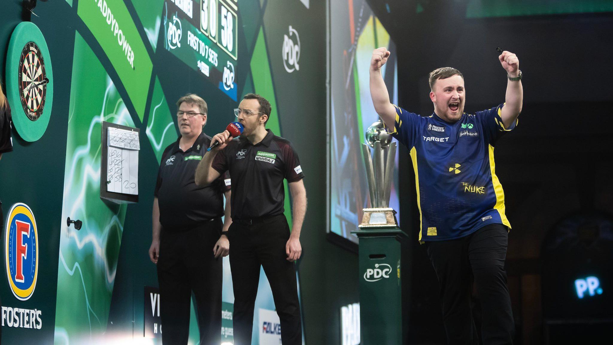 Luke Littler celebrates on the right of the image with his arms in the air fist pumping towards the crowd. On the left, Huw Ware looks at the dartboard and holds a microphone to his mouth as he is about to announce the score. Another referee is stood to the left of him. The World Championship trophy sits in the centre of the image.