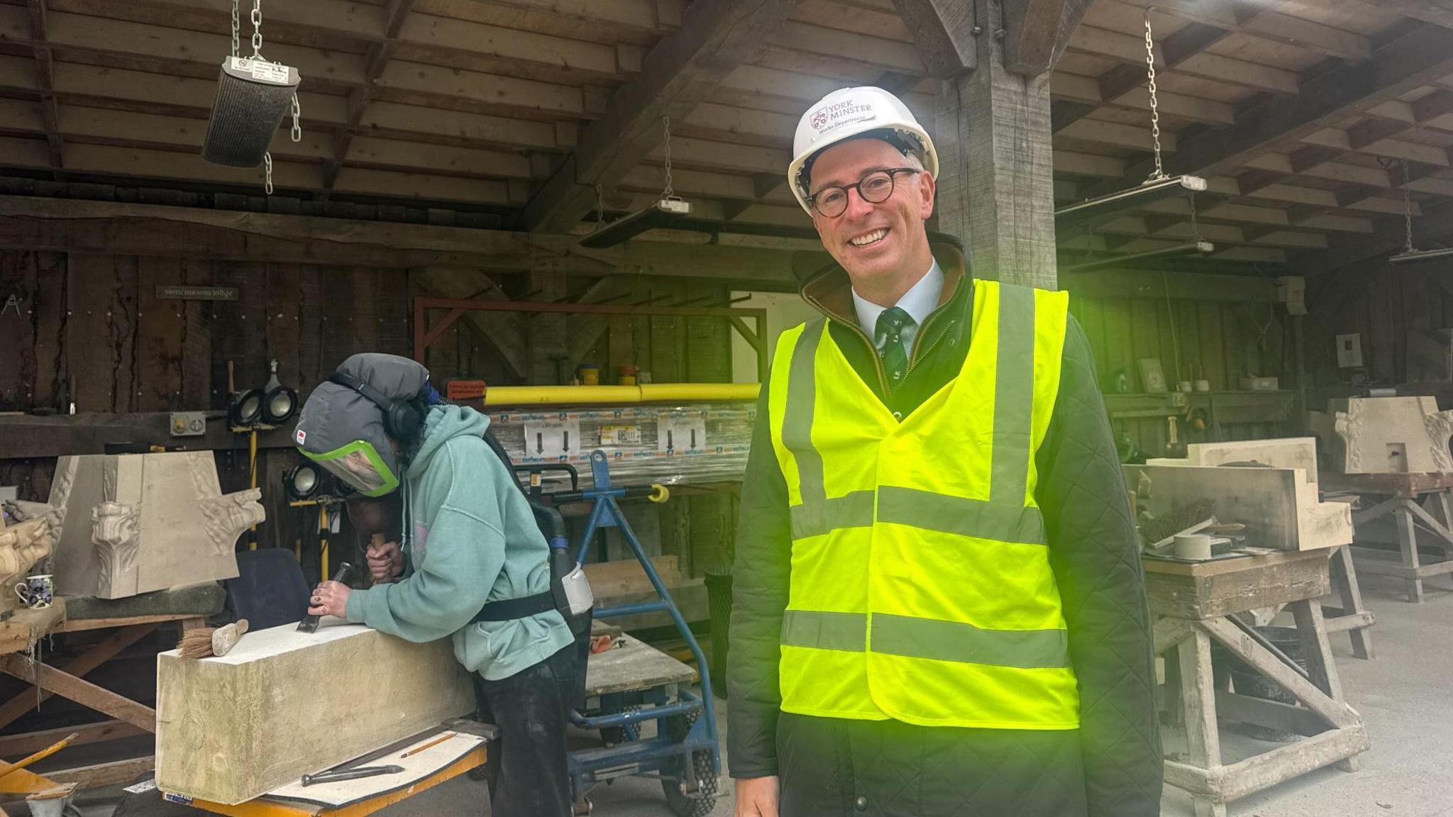 A man in a hi-vis jacket and helmet standing next to a female stonemason  chiselling a block of stone 