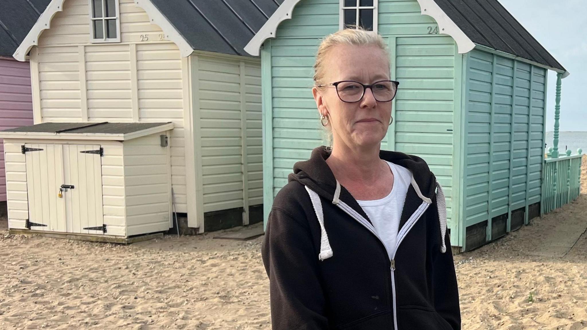 Claire Douglas has blonde hair tied back, black-framed glasses, and wears a dark-coloured hooded top, as she looks to the camera, with the sandy beach and the back of beach huts behind her.