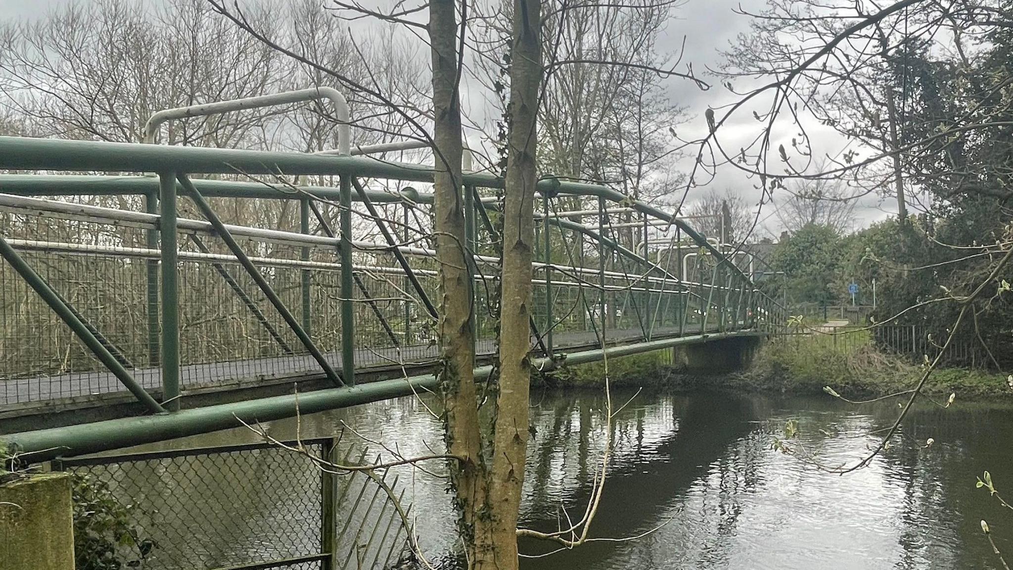 An ageing bridge, which is green with wire mesh sides going across a river on a grey day.