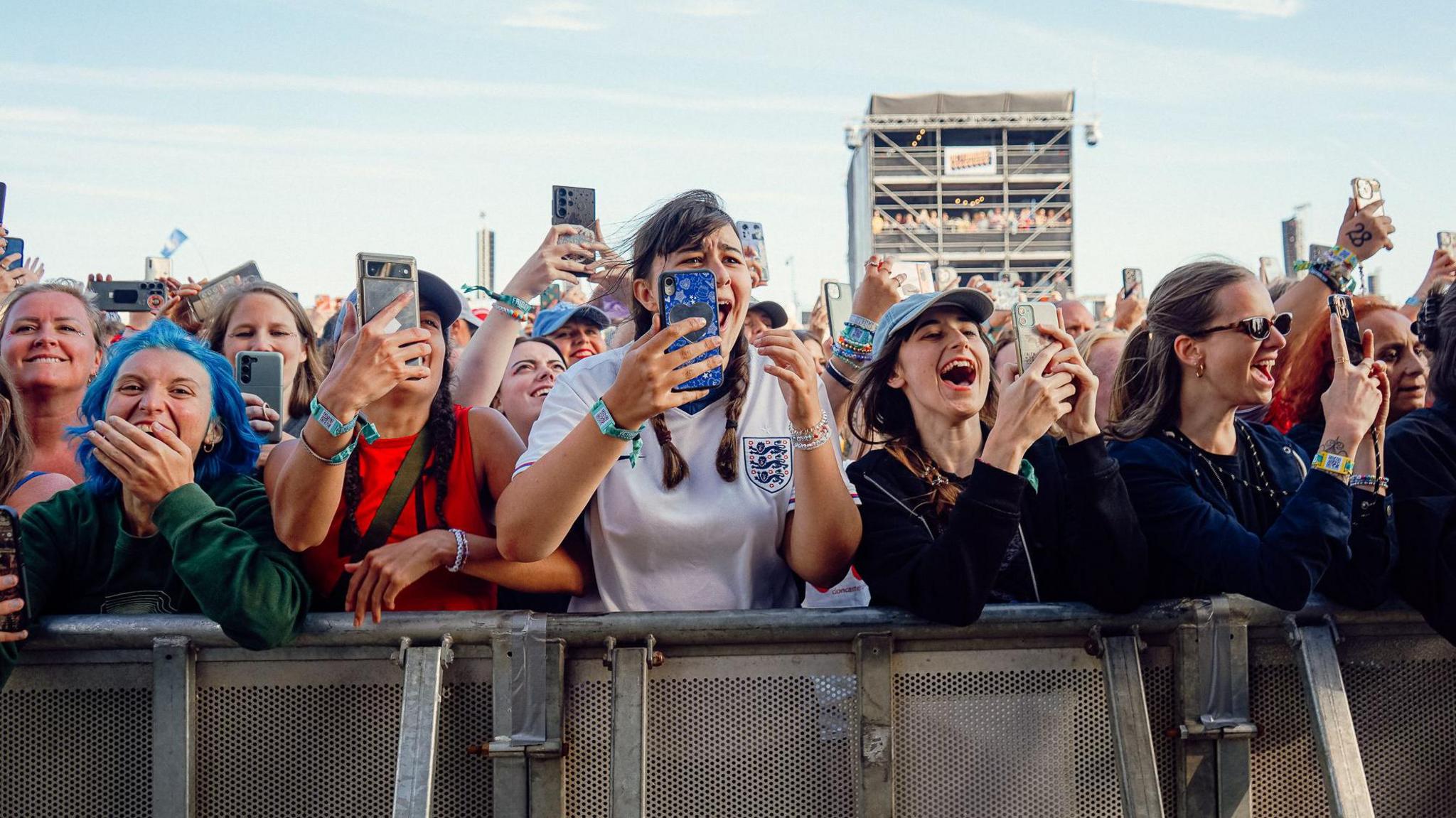 Louis Tomlinson fans at the front of the crowd at the barriers. They look very excited and most are filming on their phones. A girl in the centre wears an England shirt