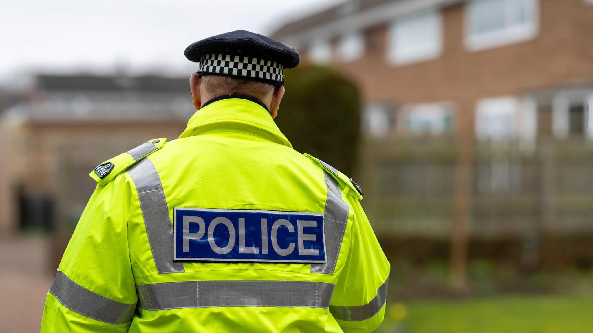 A stock image of a male police officer taken from behind to obscure his identity. He wears a hi-vis jacket and black hat.