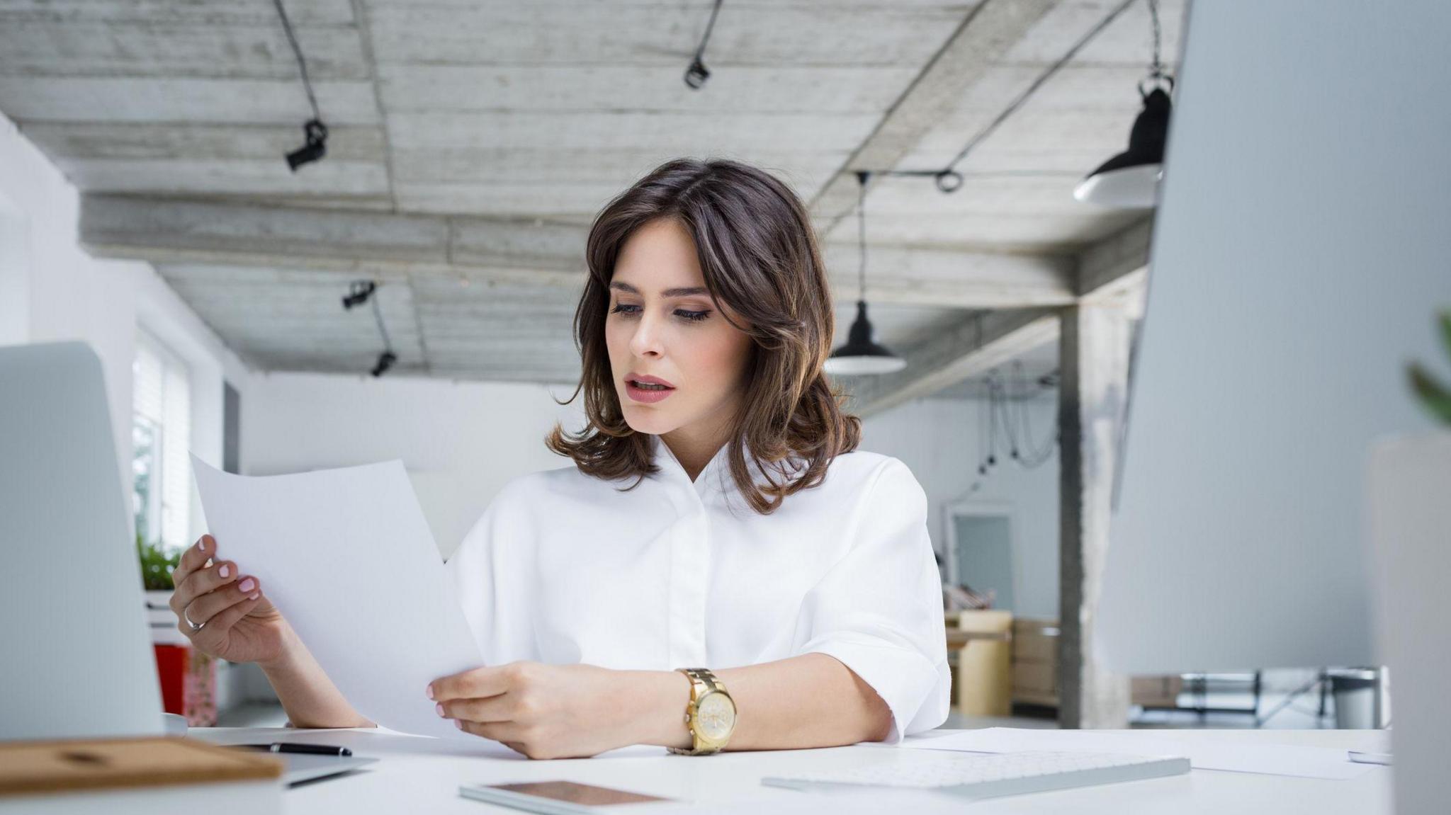 A woman sits at a desk in a very clean and white office. She is reading a piece of paper with a concerned look on her face. Her hair is brown and she is wearing a white shirt.