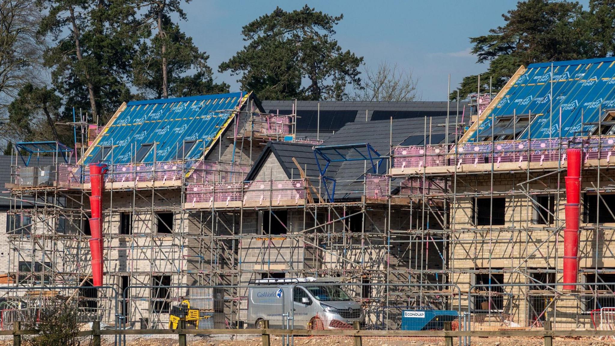 Scaffolding surrounds a couple of new build houses under construction. A silver van is parked outside. Three trees can be seen behind them on what is a sunny day.