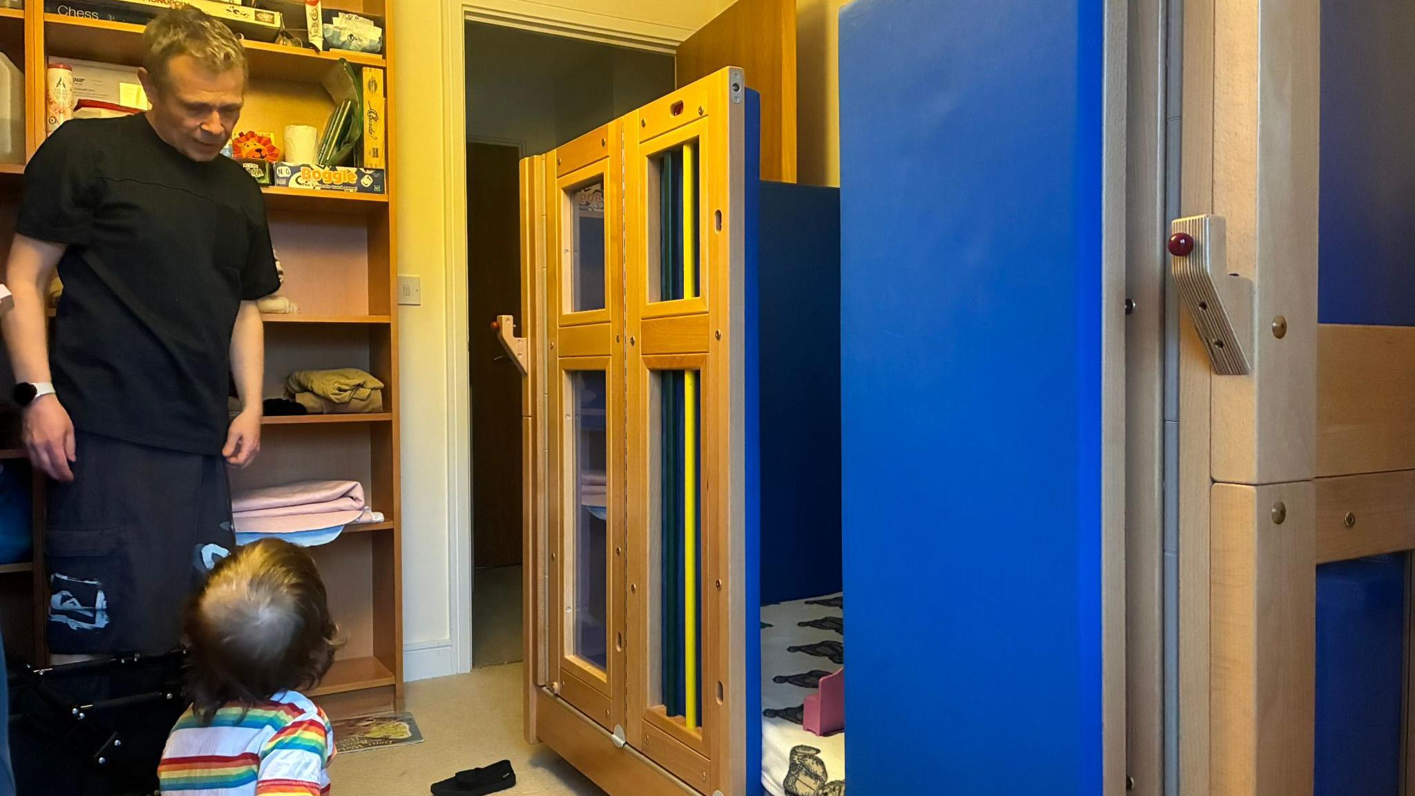 A man and young boy in a bedroom. Next to them is a large wooden structure which is used as a bed. It has blue padded sides. The man is standing. The boy is sitting.