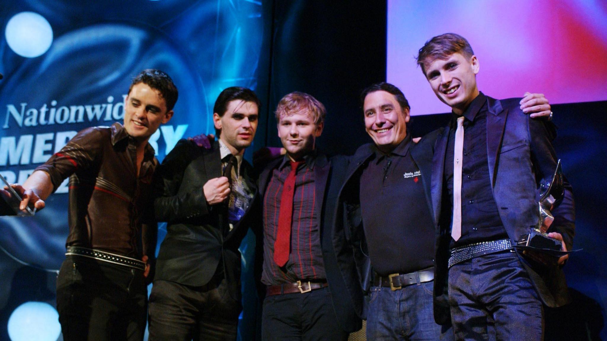 Host Jools Holland congratulates Franz Ferdinand on winning the Mercury Music Prize in 2004. The band and Holland have their arms wrapped around each other while singer Alex Kapranos, on the far right, holds the award in his hand. The band are mostly dressed suits with ties, except for guitarist Nick McCarthy who is dressed casually.