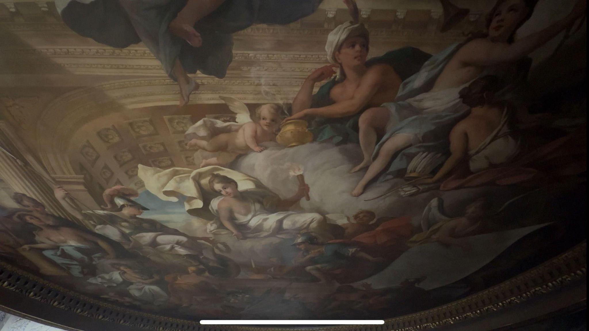 A wide image of a large baroque painting on the ceiling of the great hall in Blenheim Palace, depicting several historical figures in a grand ballroom