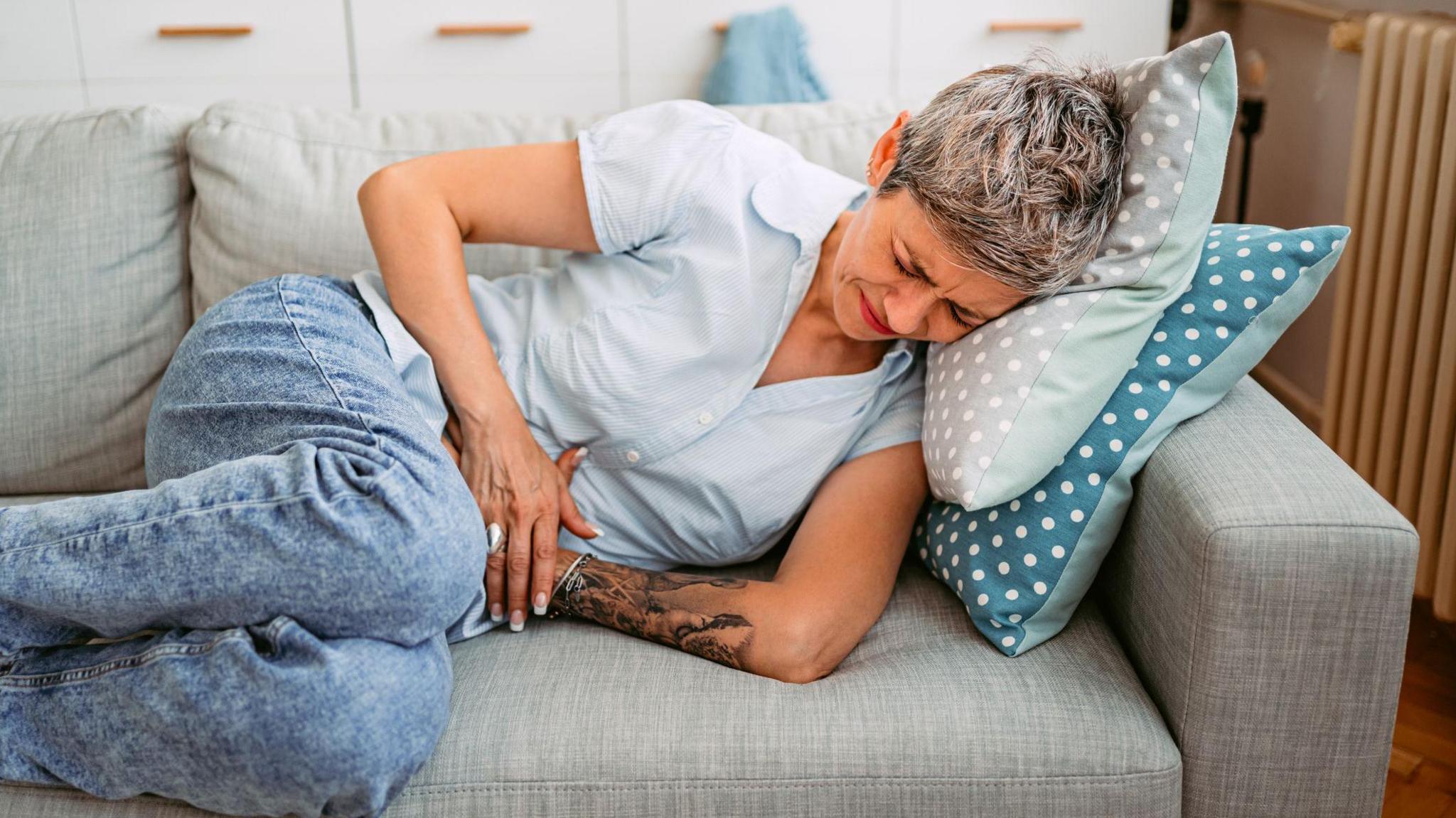 A woman with short hair lies on a sofa, clutching her stomach. She has short grey hair and wears a blue shirt with blue jeans. 

The sofa is grey. 