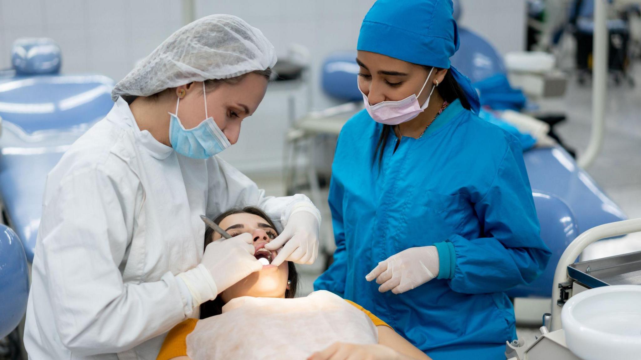 Two female dentists carry out an examination on a third woman's mouth. They are wearing blue and white PPE. The patient is lying down with her mouth open.