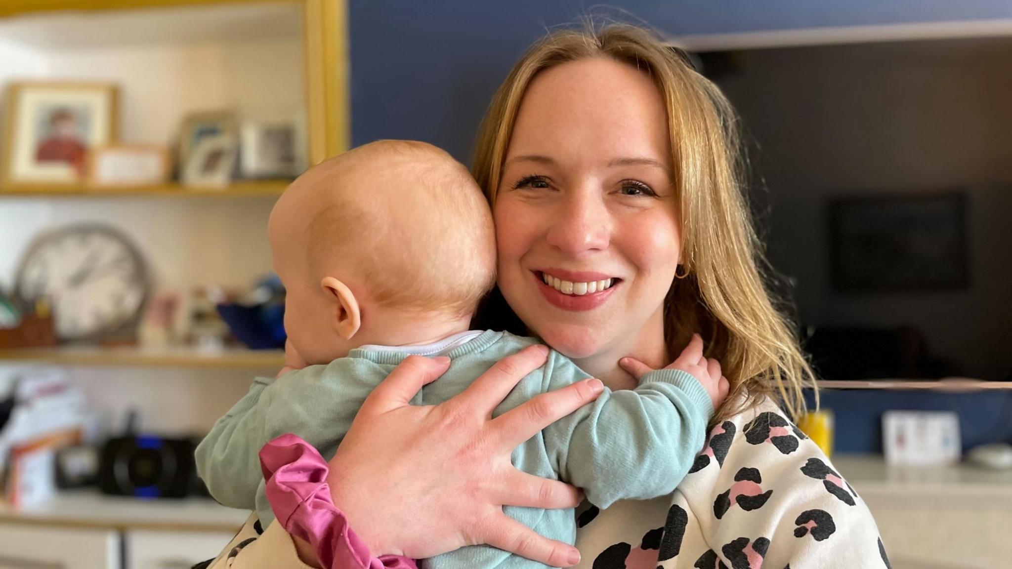 A photo of Sophie with baby Nora. Sophie looks directly at the camera. She has long blonde hair. Nora is being held up with her back to the camera.