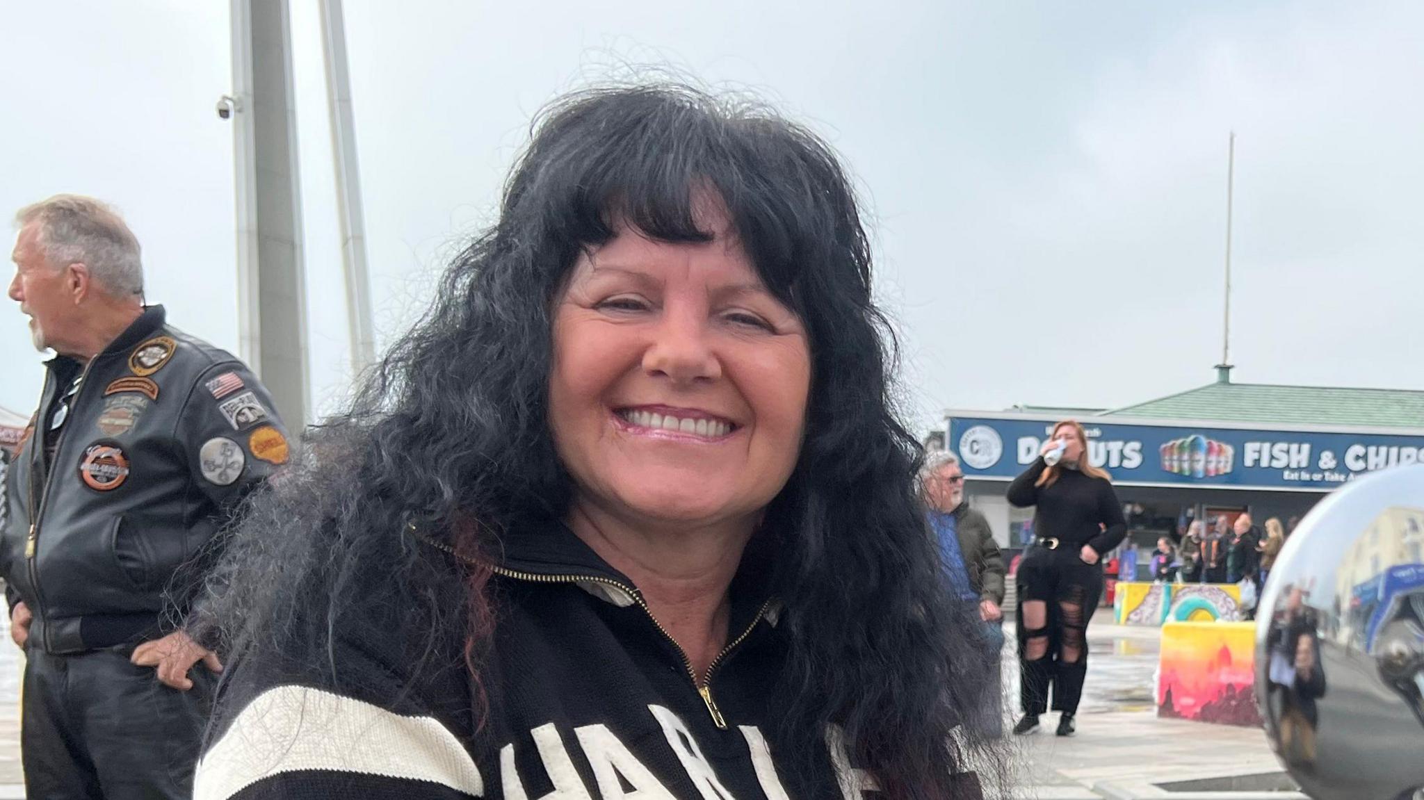Karen Gelsthorpe smiles at the camera as she sits on her Harley-Davidson motorbike. She has shoulder length dark hair and wears a black jumper with the words Harley Davidson written on the front, the jumper's arms are white and black striped.