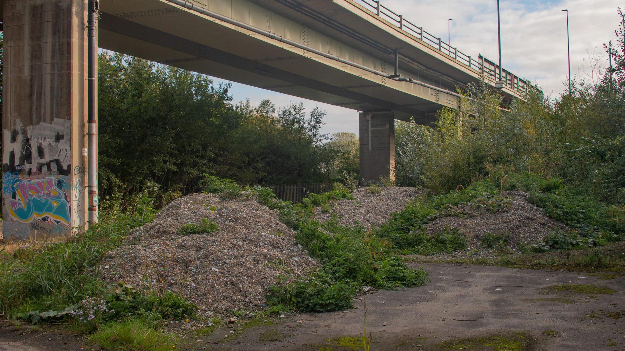 Large piles of waste under a motorway bridge with green vegetation sprouting from them