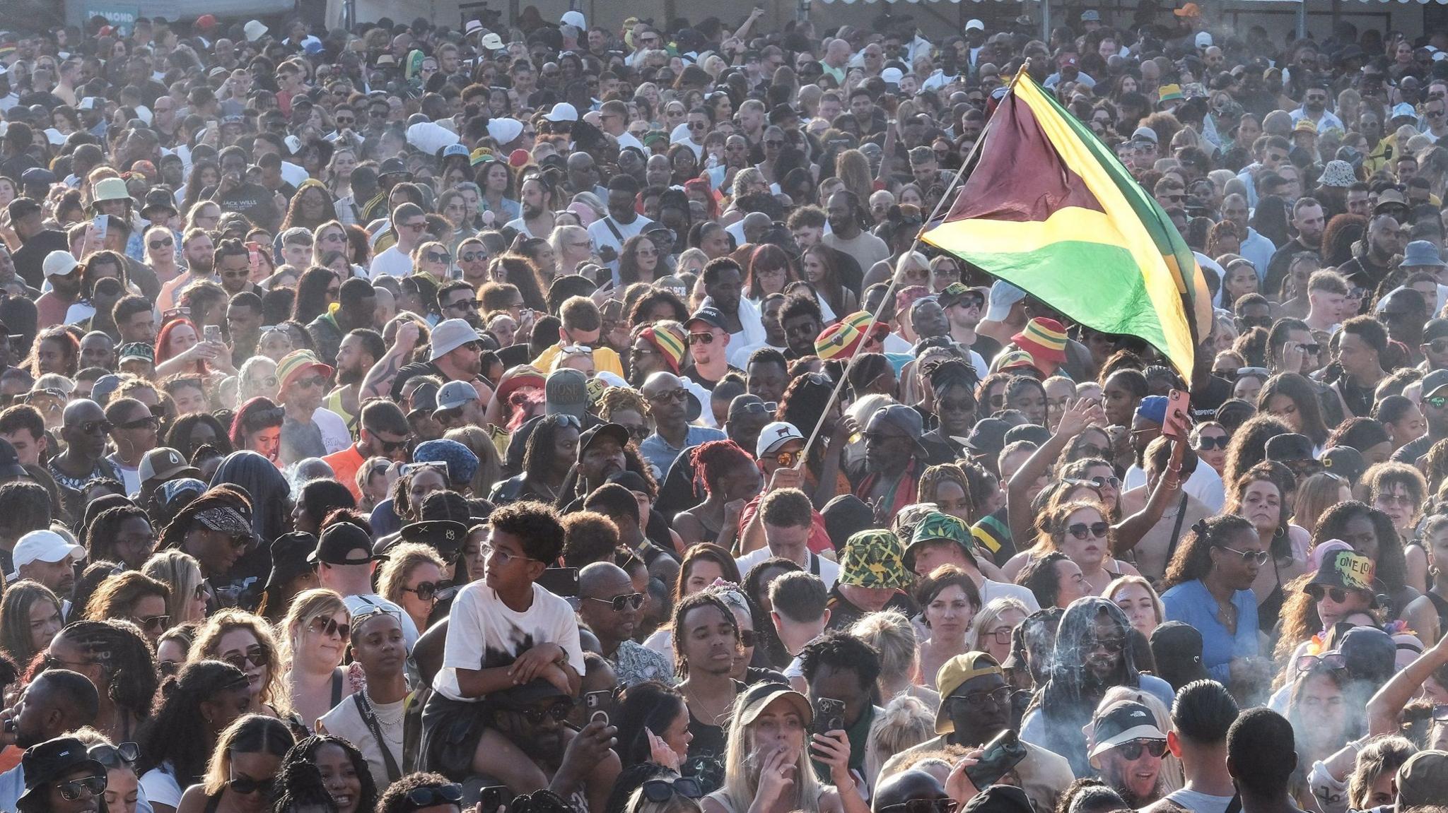 A huge crowd of people at a festival. A Jamaican flag is flown prominently.