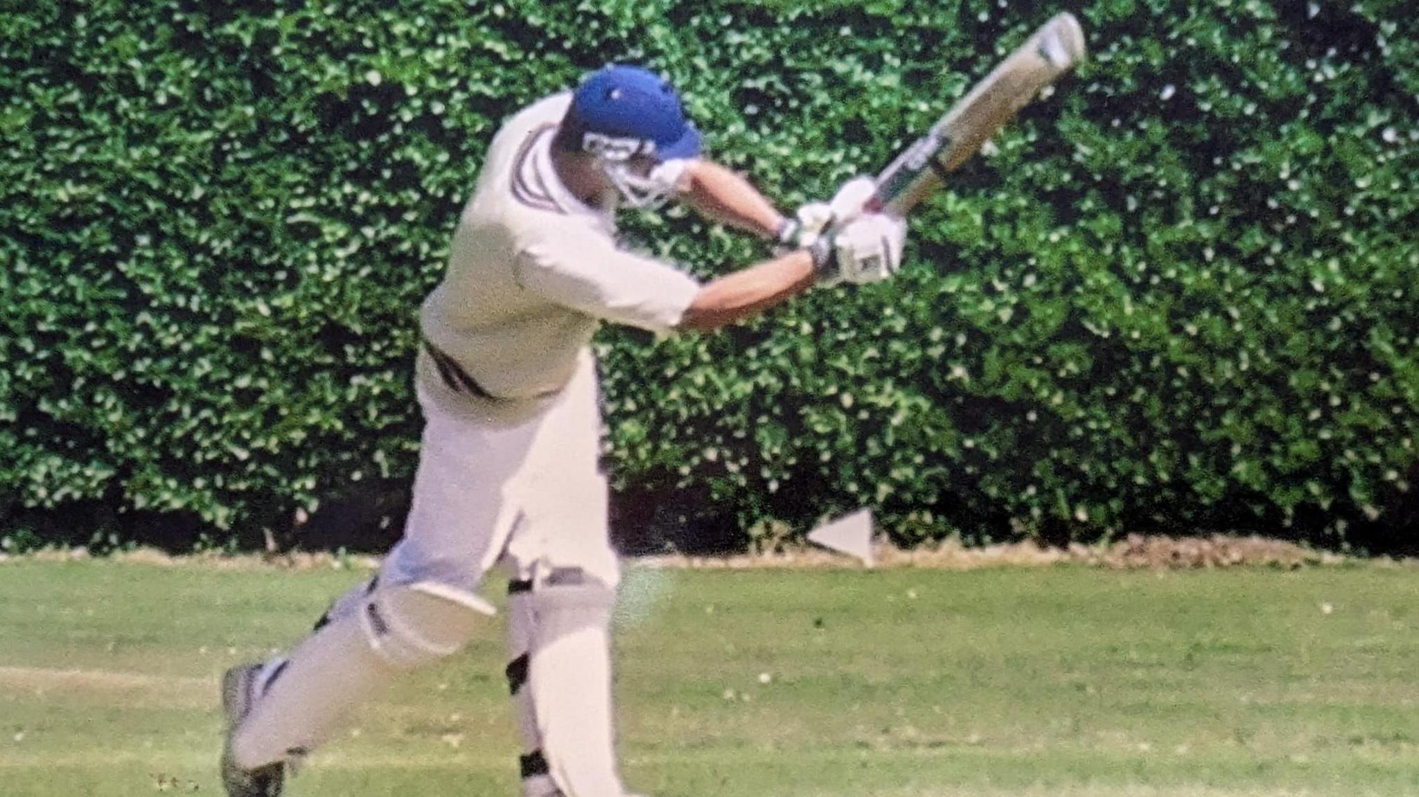 George Rudd in cricket whites and a blue helmet, swinging his cricket bat up as if he has just hit a ball, with a hedge behind him