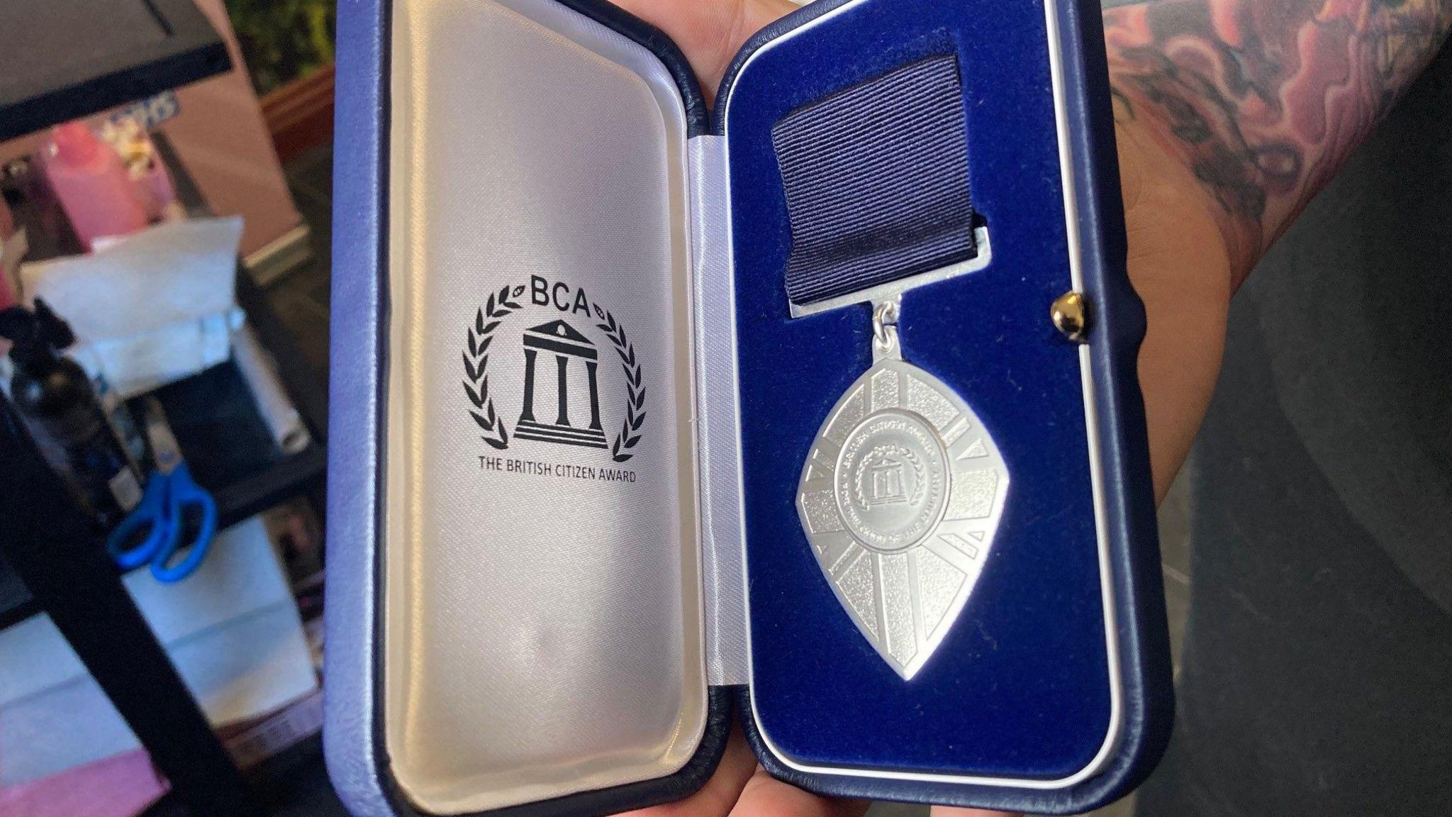 A silver medal with blue ribbon in a dark blue presentation box with a the British Citizen Award logo on a white silver liner.