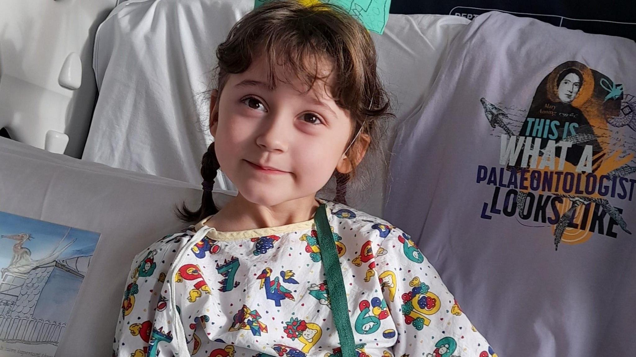 Sylvie during her treatment on Bellhouse Ward. She is looking away from the camera. She is wearing a white pijalams with various colourful characters on. Her hair is braided. She is lying on several cushions.