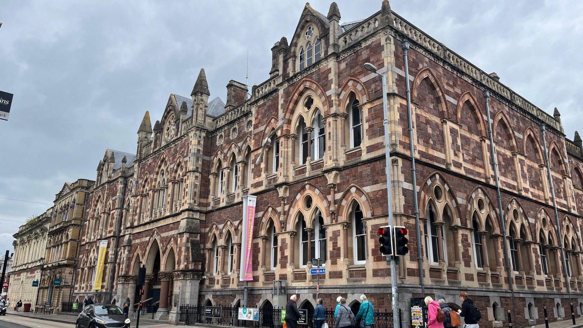 A picture of outside Exeter museum. It has red brick and has arches around its windows. There are two flags near the entrance and there are people walking outside of it.