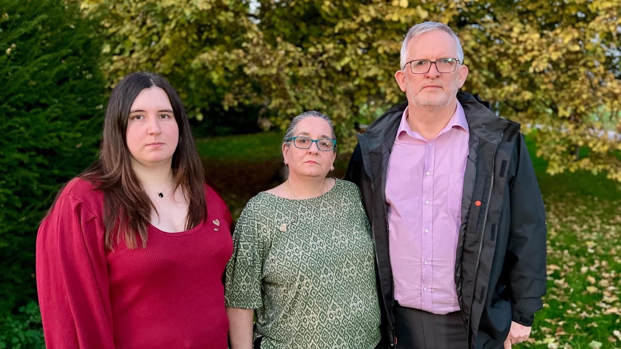 Declan's sister Kaitlyn, mother Sam and father Graeme, standing together in front of trees shedding their leaves. Kaitlyn is in her 30s and is wearing a red jumper, while Declan's mother is wearing a patterned green top and has blue rimmed glasses. His father has grey hair, glasses and a light beard and is wearing a black rain jacket, a purple shirt and grey suit trousers. They are looking at the camera seriously.