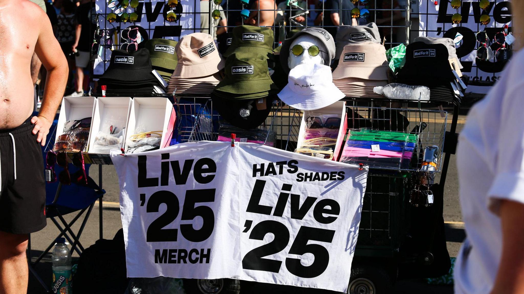 A bare-chested man stands next to a stall selling black, green and beige bucket hats and various sunglasses. It has a sign on it which reads: Live '25 Merch Hats Shades
