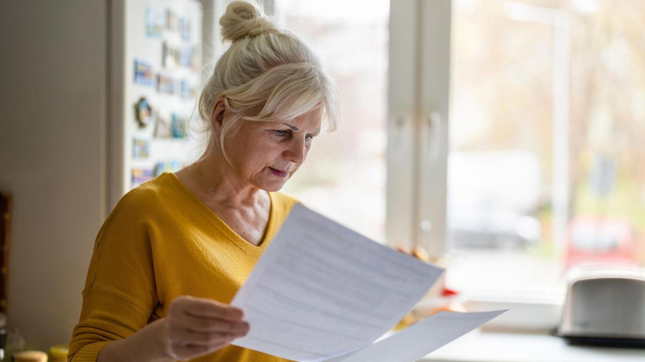 Woman looks at paperwork in a kitchen with a fridge-freezer and a toaster behind her.