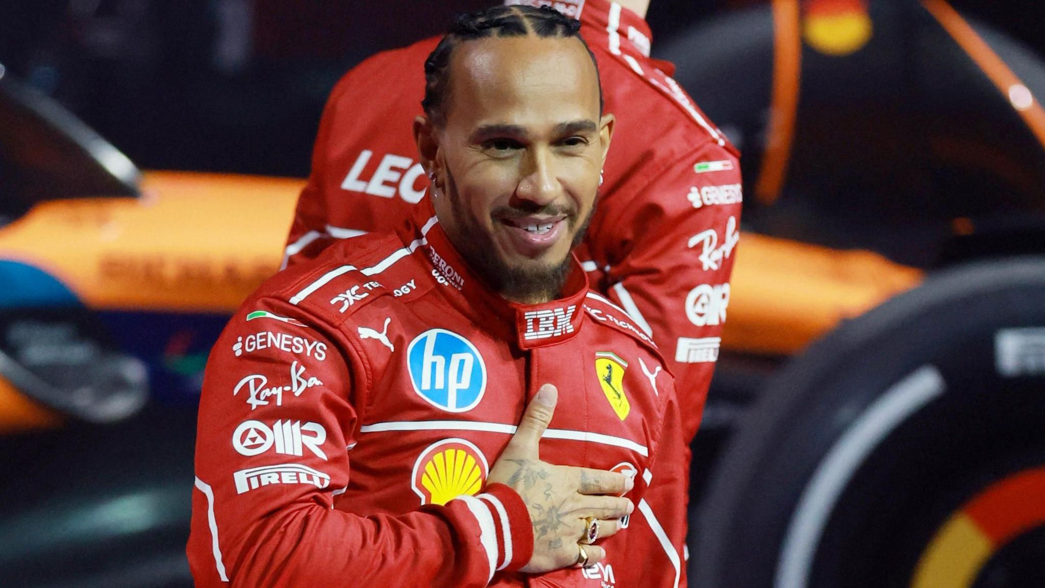 Lewis Hamilton holds his right hand across his chest, just below the Ferrari badge, while wearing a race uniform at the F1 75 season launch event at the O2 Arena